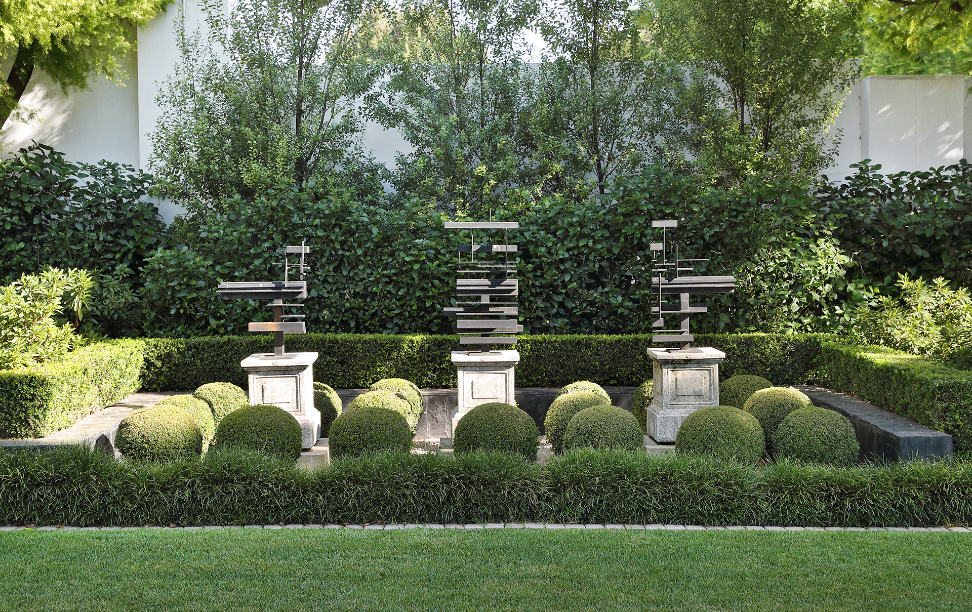 Formal garden with hedges and three modern metal sculptures on plinths, by Young Garden Design.